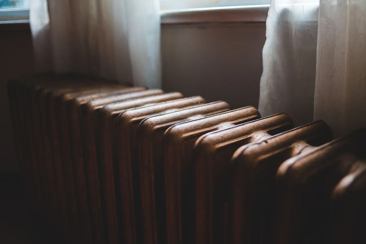 Aged heating battery made of cast iron near windowsill with curtains in house