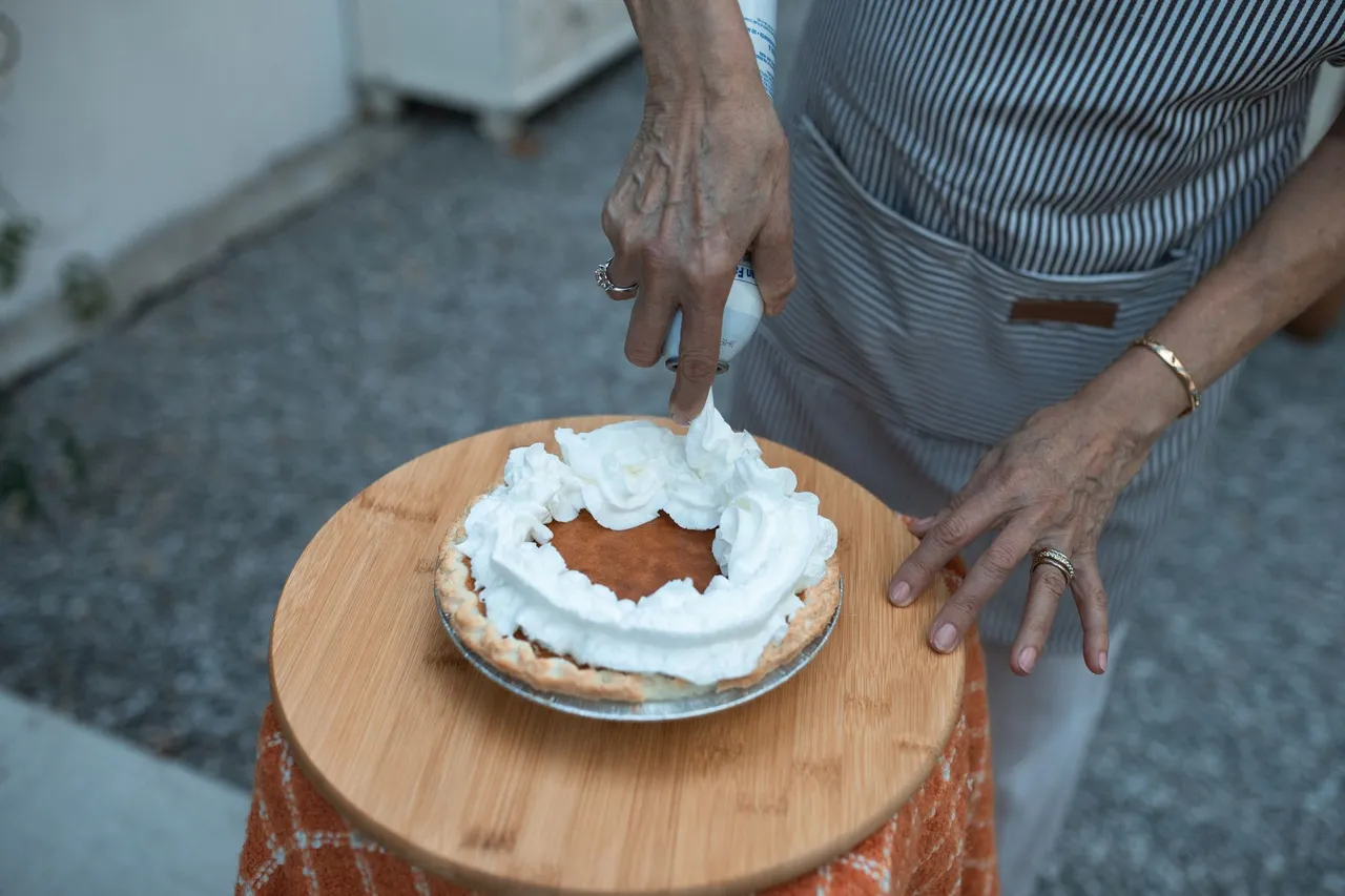 Close-up of hands adding whipped cream to a delicious pumpkin pie on a wooden plate.