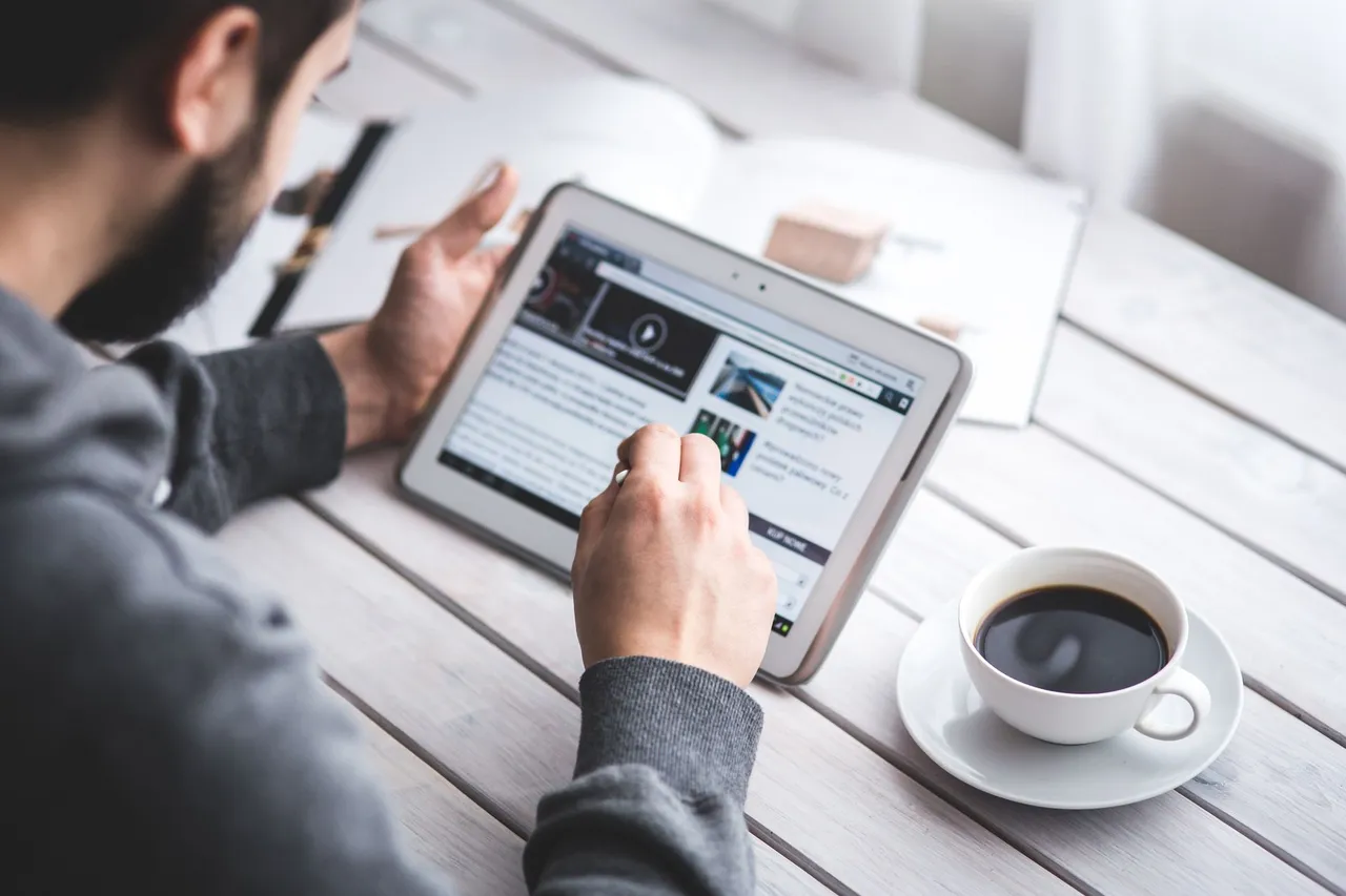 Man browsing the news on a tablet while holding a stylus, with a cup of coffee.