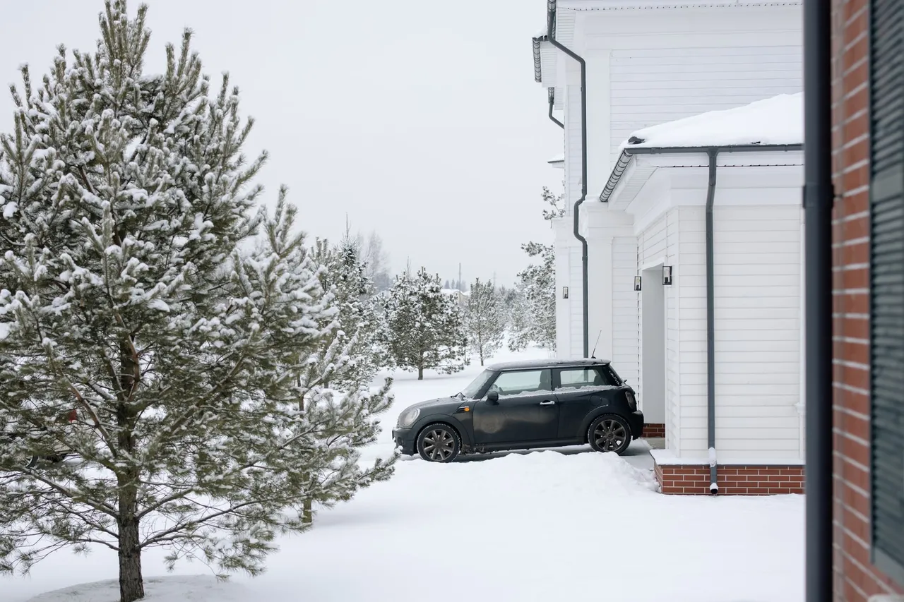 A Mini Cooper parked in a snowy driveway beside a white house during winter.