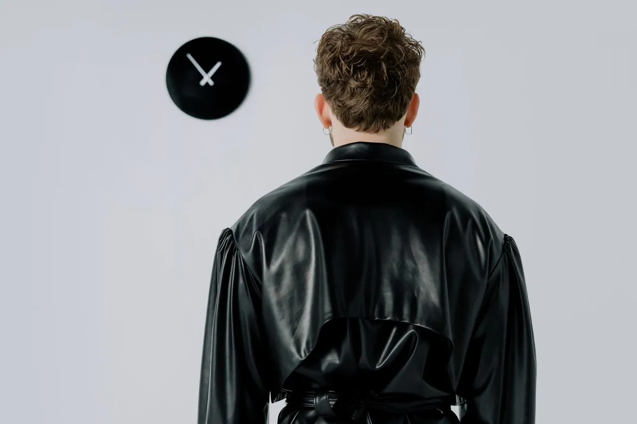 Back view of a man in a black leather jacket facing a wall clock indoors.
