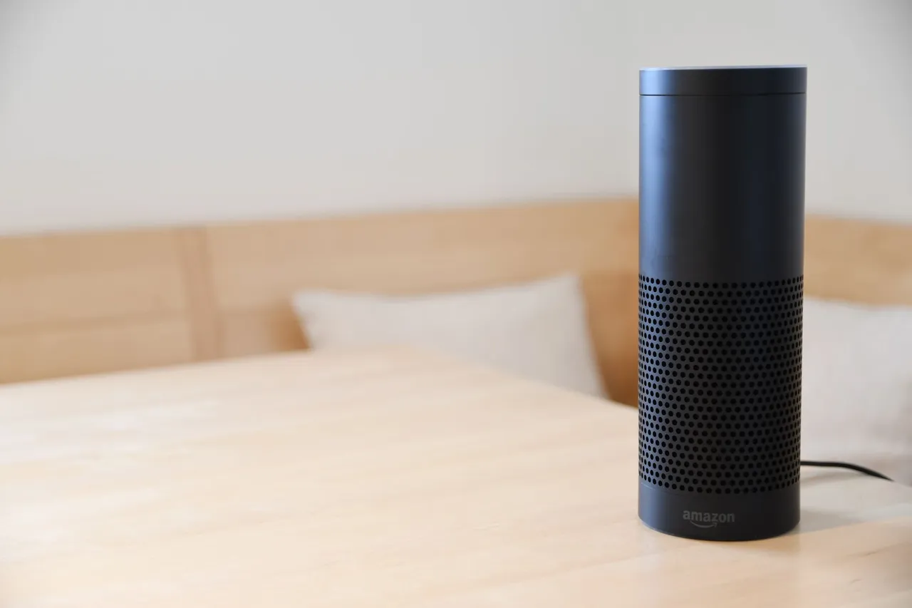 A black smart speaker resting on a light-colored wooden table in a cozy indoor setting.